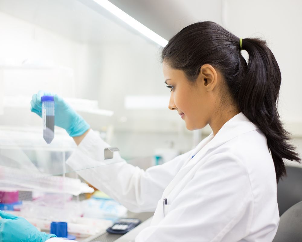A scientist in a lab coat and gloves examines a test tube at a laboratory workstation, working under a fume hood.