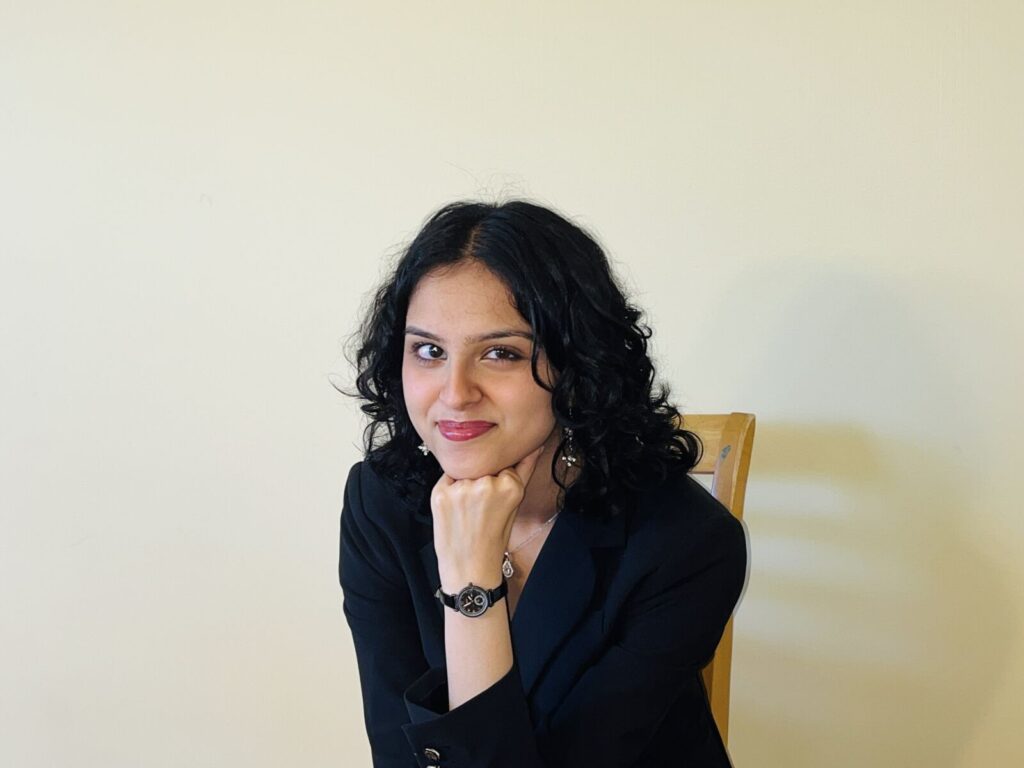 Woman with curly dark hair wearing a black blazer sits on a wooden chair, resting her chin on her fist, against a plain light-colored wall.