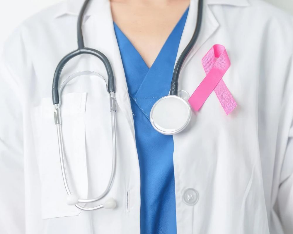 A healthcare professional wears a white coat with a stethoscope and a pink breast cancer awareness ribbon pinned to the chest.
