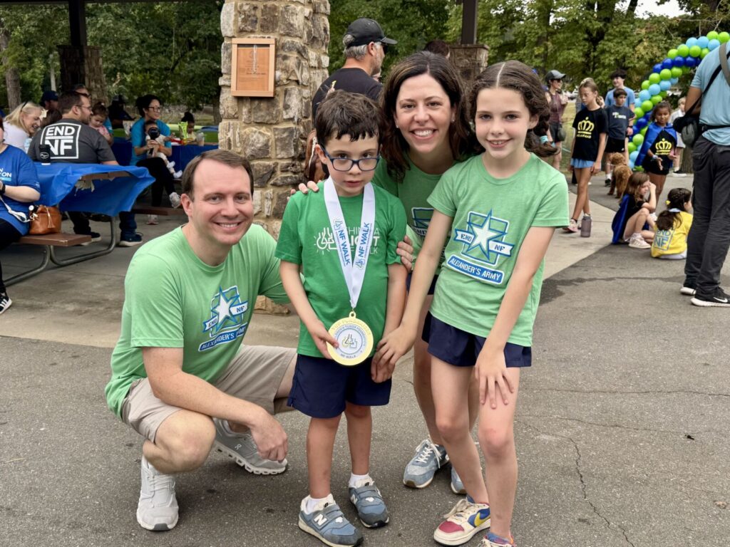A family of four wearing matching “Alexander’s Army” green t-shirts at an outdoor event, with the young boy in the center wearing a medal.